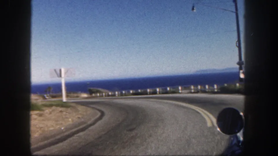 San Diego coastal road footage from 1962 shows a right-hand curve above the ocean with guardrail posts, roadside sign, and vehicle mirror.