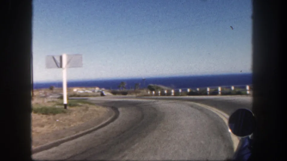A 1962 coastal driving frame shows a large white roadside sign near a curving road, with the ocean horizon and vehicle mirror visible.