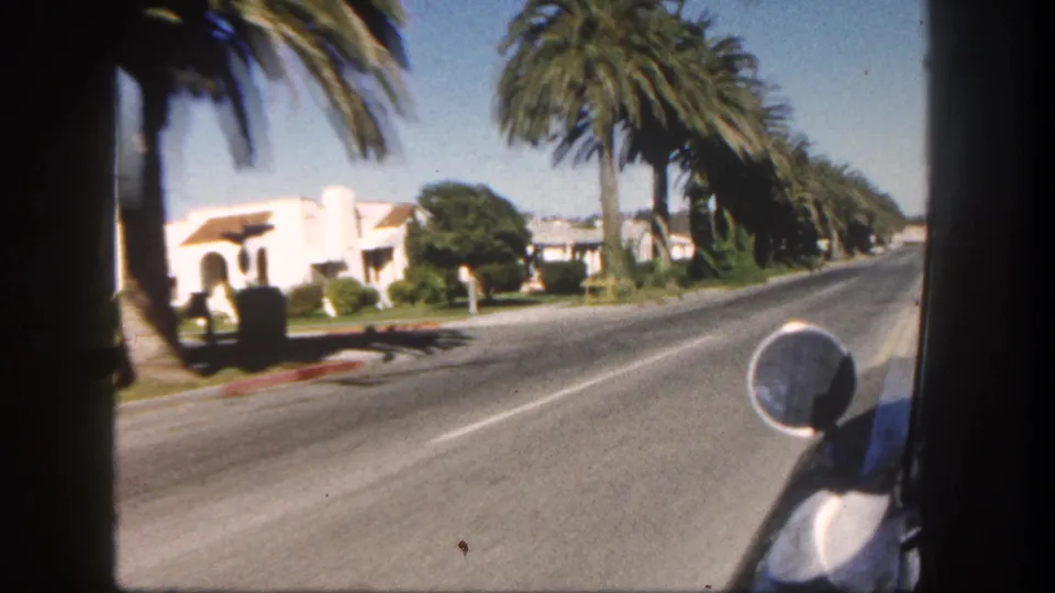 Forward-moving 1962 San Diego street view showing palms, white residential buildings, lawns, and the car mirror near the right edge.
