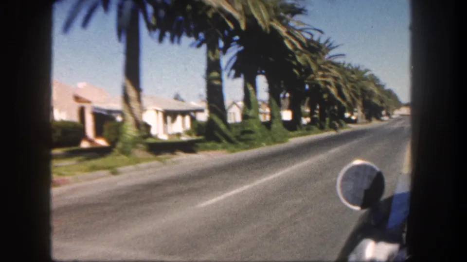 End-frame view from a moving car on a 1962 San Diego palm-lined residential street with stucco homes and a side mirror visible.