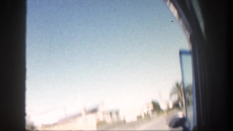 1962 San Diego home-movie frame from a car window, showing palms, road, and bright residential exteriors.