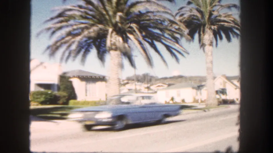 A light blue mid-century car passes through a 1962 San Diego residential street scene with palms and houses.