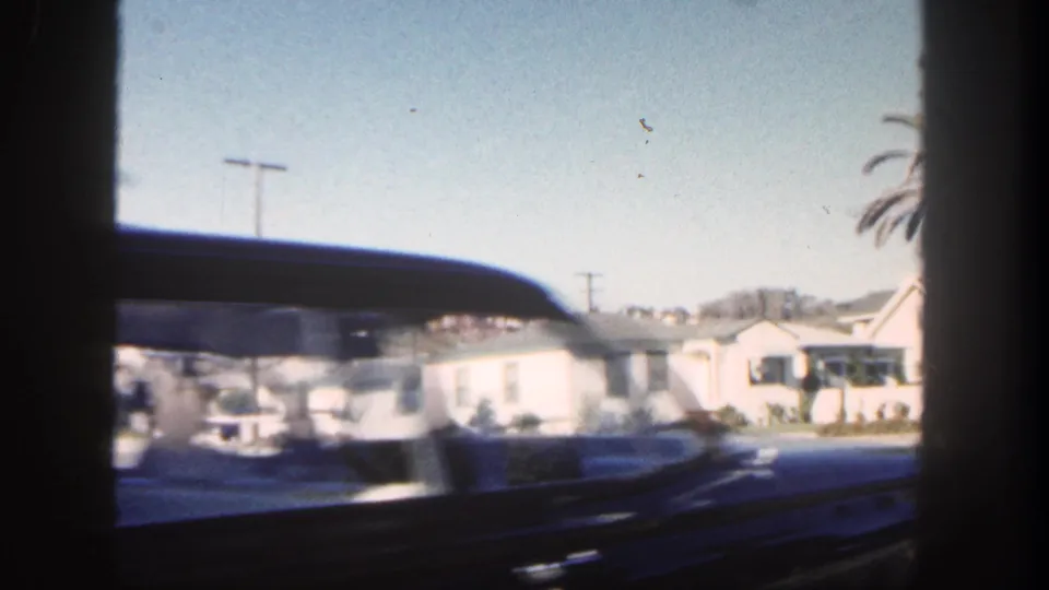 Moving 8mm view of a dark car in the foreground with white San Diego houses, palms, and utility poles behind it.