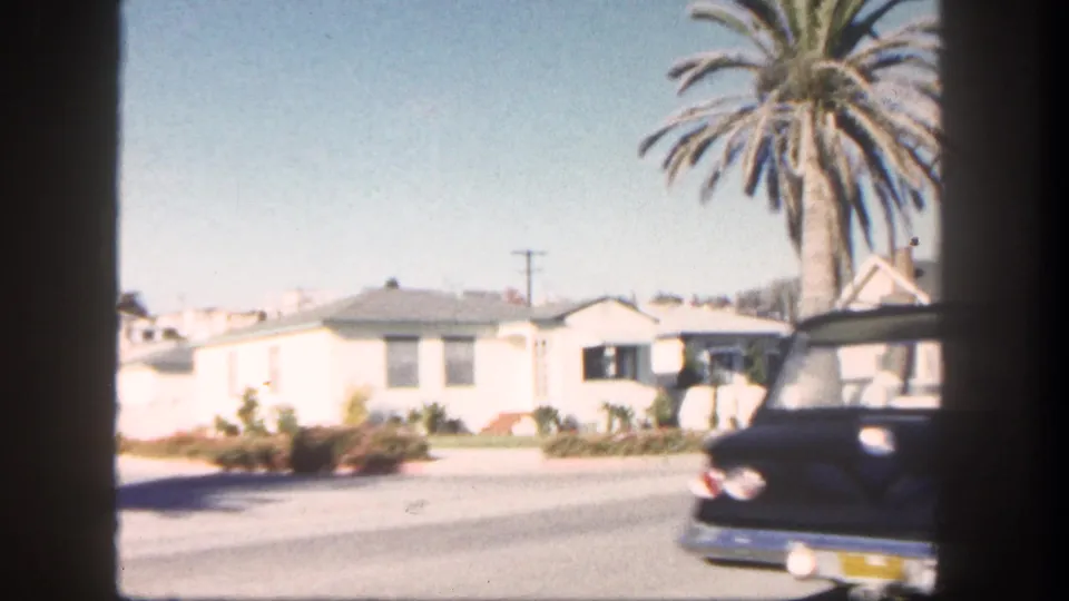 1962 San Diego street view with white houses, a tall palm, planted yards, and a dark car at the frame edge.