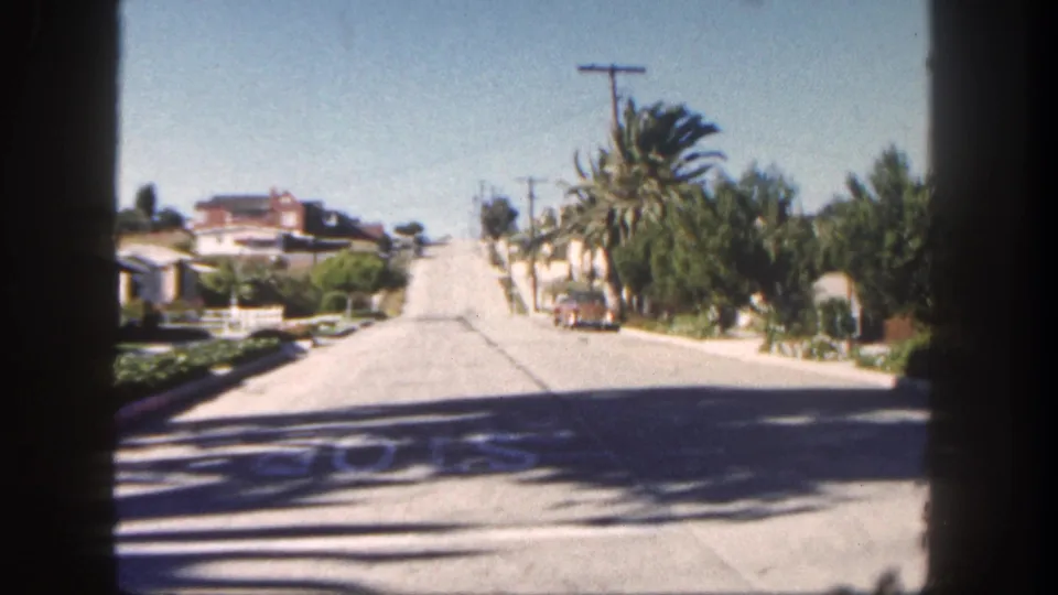 1962 San Diego residential street with palm trees, low houses, utility poles, and bright sun, filmed from behind window bars.