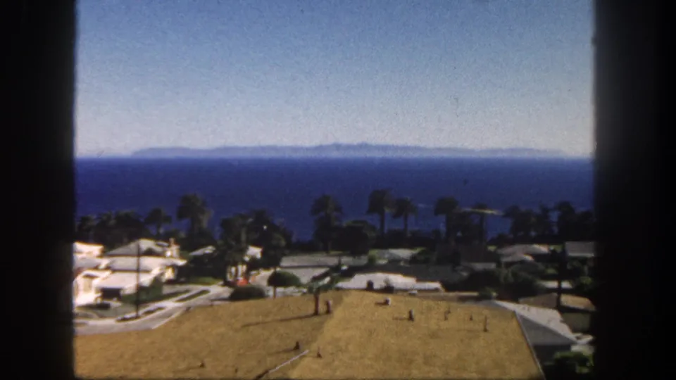 1962 San Diego coastal view framed by a dark window edge, with palm trees, low houses, and the ocean beyond.