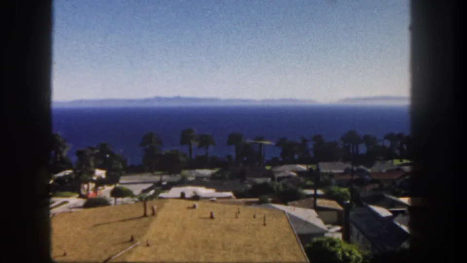 1962 San Diego home-movie still of a coastal neighborhood, palm trees, rooftops, and the Pacific Ocean in the distance.