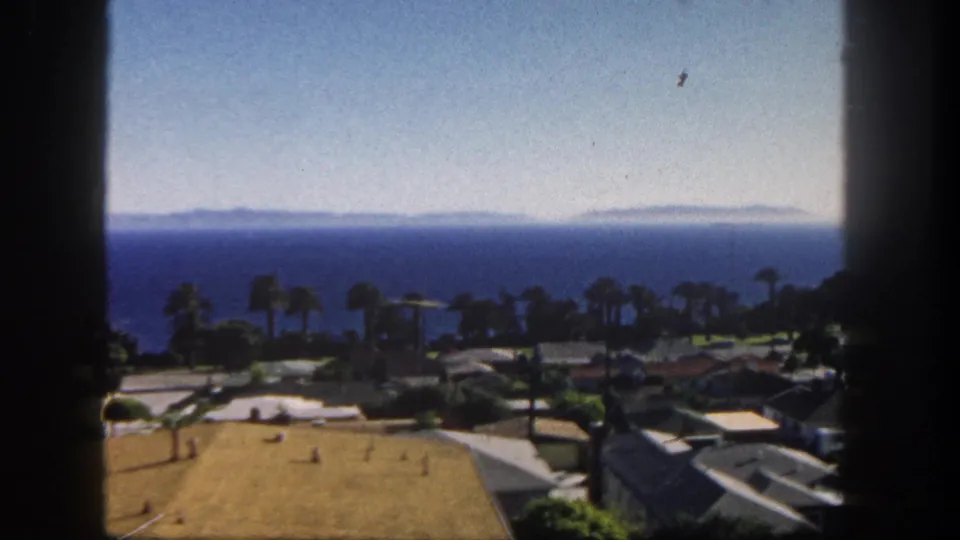 1962 San Diego home-movie frame showing a palm-lined coastal neighborhood, low rooftops, dry lawn, blue water, and distant shoreline.