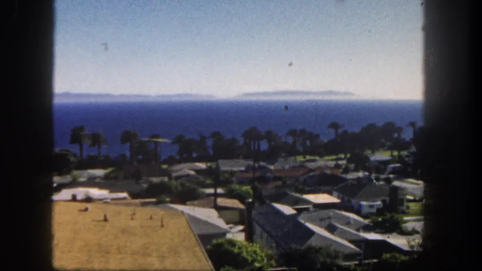 1962 San Diego coastal residential view with palm trees, low homes, rooftops, blue ocean, distant land, and dark window-like edges.