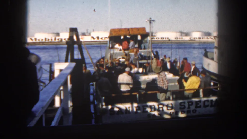 The home-movie frame shows men gathered at a docked San Diego charter boat, with harbor water and Mobil Oil storage tanks across the channel.