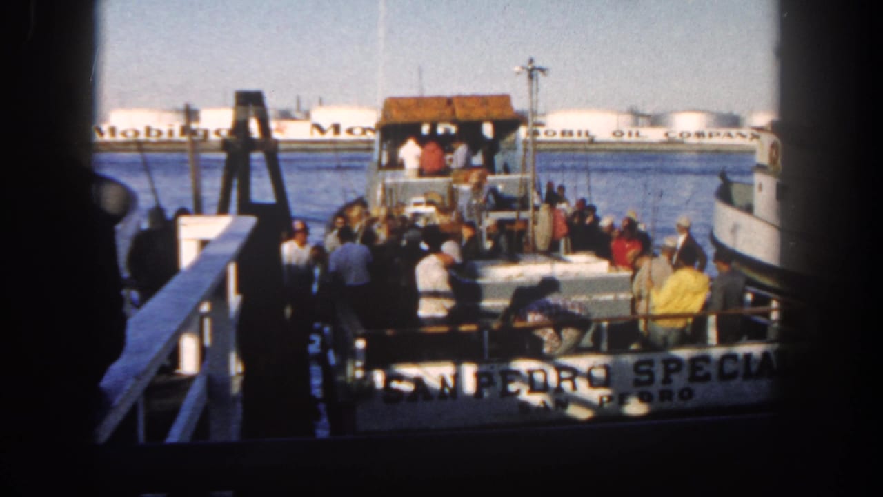 Crowded 1962 San Diego ferry deck viewed from the dock, with people clustered near the cabin and Mobil Oil tanks behind.
