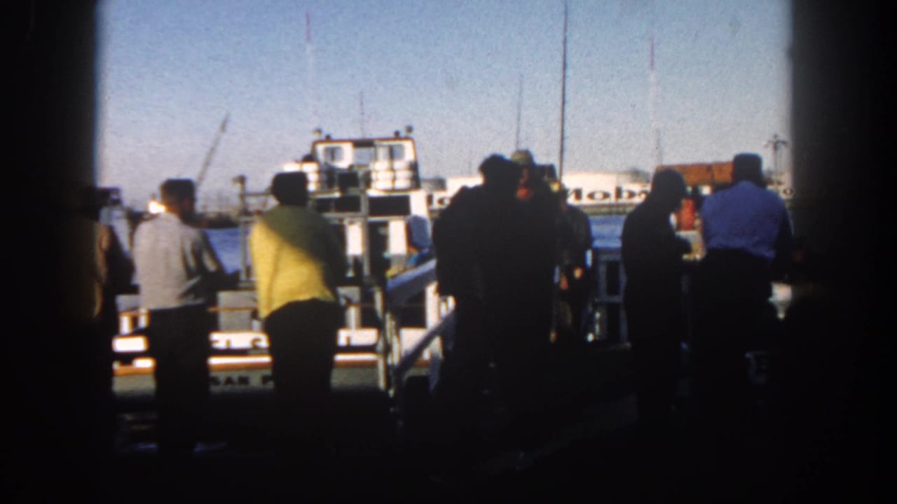 Passengers in mid-century clothing stand along a San Diego dock facing a white harbor vessel, with industrial tanks visible across the water.