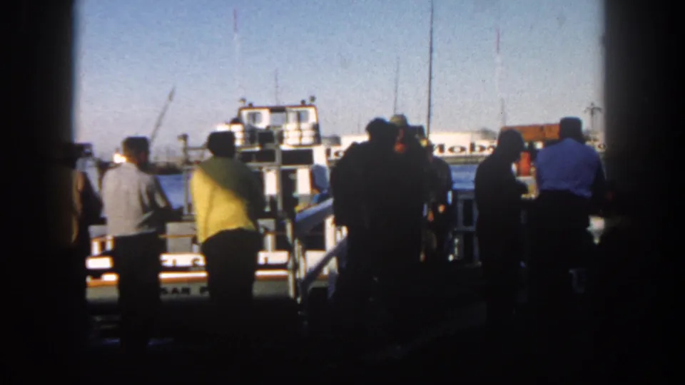 Passengers in mid-century clothing stand along a San Diego dock facing a white harbor vessel, with industrial tanks visible across the water.