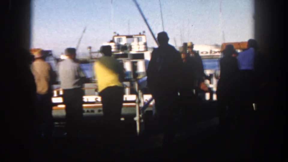 The home-movie frame shows a shaded dock crowd in San Diego looking toward a white harbor boat, with cranes and oil tanks in the distance.