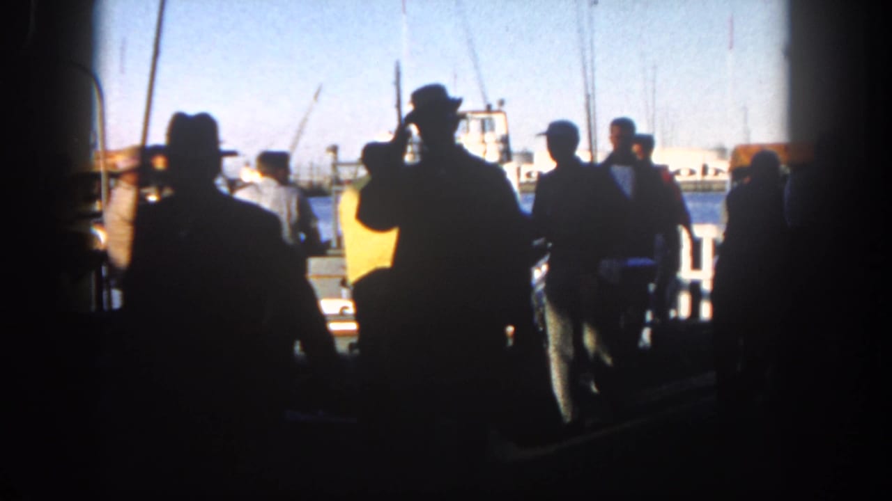 Men appear in silhouette along a San Diego dock in 1962, with moored boats, water, and masts lit by afternoon sun behind them.