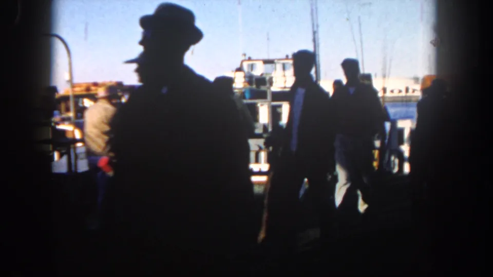 Two adult men pass through the shaded foreground of a 1962 San Diego harbor shot, with boats and dock railings visible beyond.