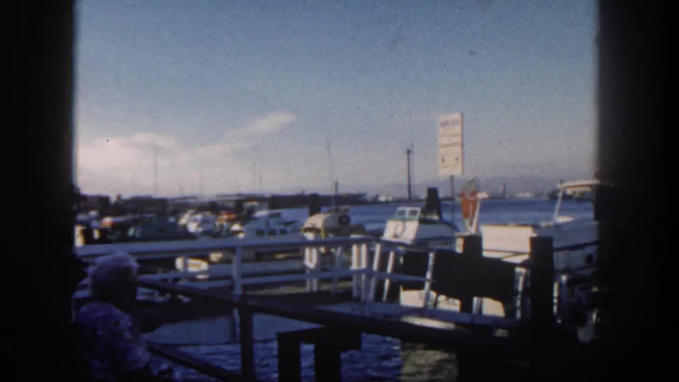 San Diego harbor scene from 1962 showing boats, parked cars, dock railings, a sign, water, and a seated adult in the shaded foreground.