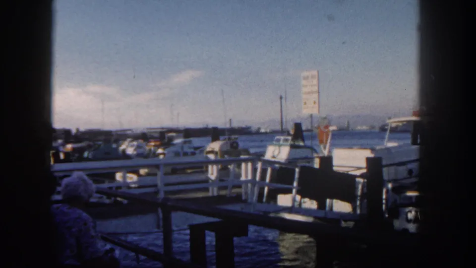 1962 San Diego harbor scene with small boats tied near a pier, a sign shape in the distance, and a seated person partly visible in shade.
