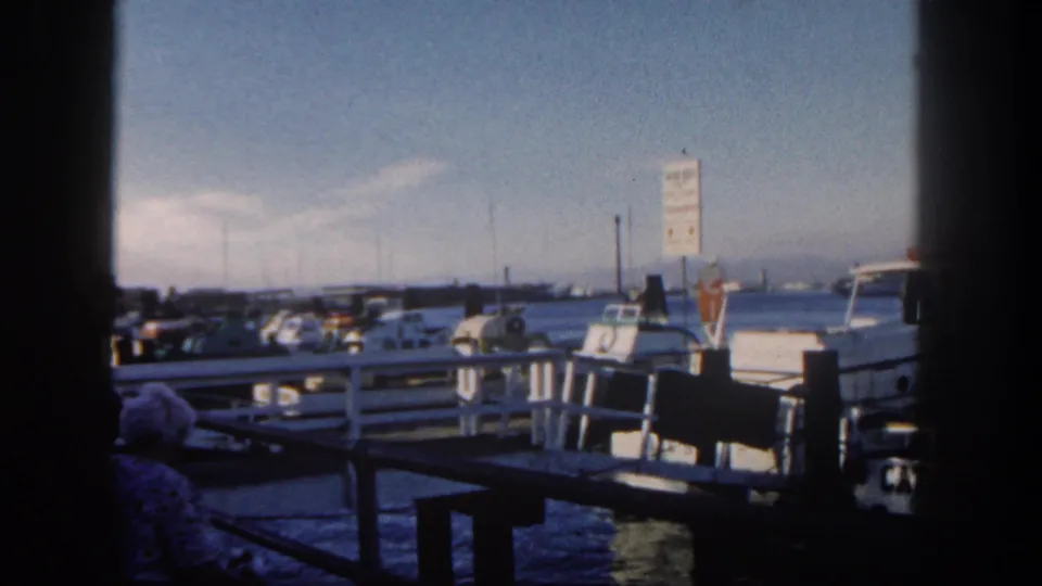 A 1962 San Diego marina view shows docked pleasure boats, wooden pier rails, water, and a white-haired person seated near the camera.