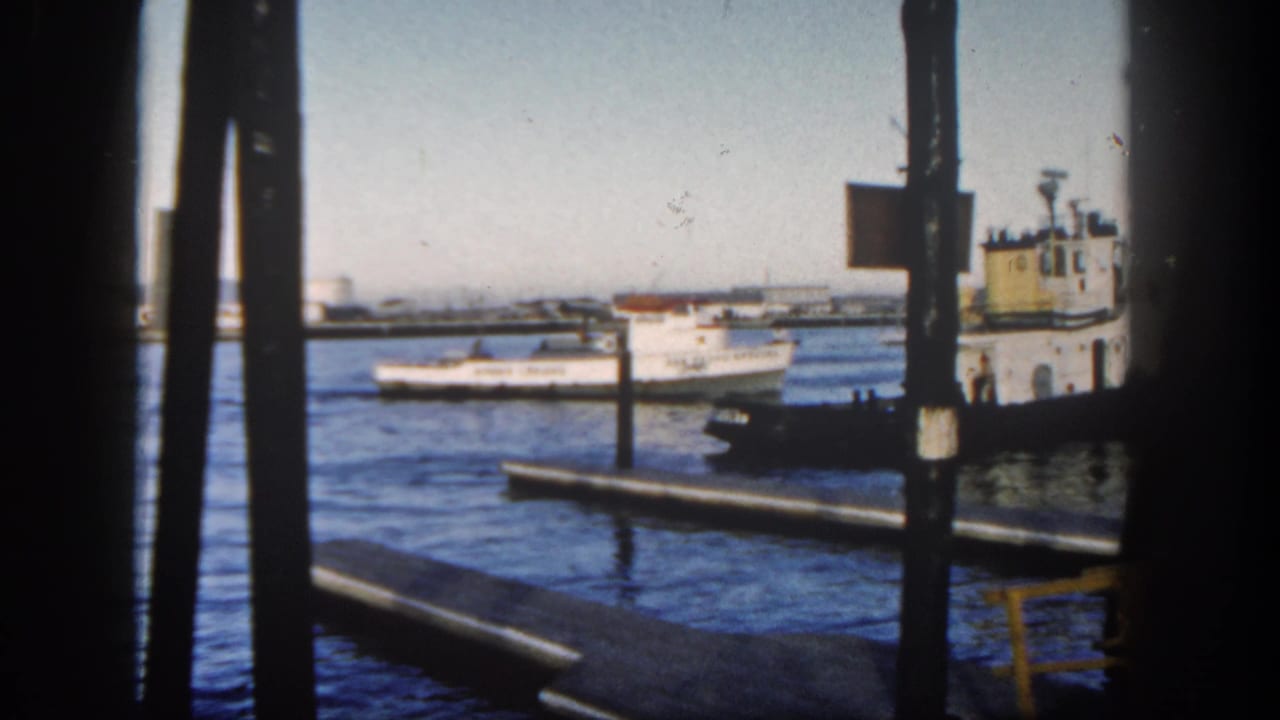White fishing boat moves across harbor water near pier posts and a darker vessel.