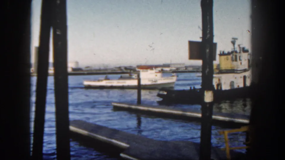 A white fishing boat passes through the San Diego harbor frame in 1962, partly obscured by dark pier posts with dock edges and another vessel nearby.