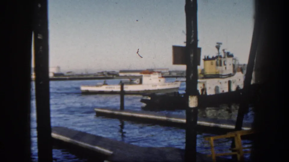 The harbor view shows a white fishing boat near a darker vessel and wooden dock structures, framed by vertical pier supports in the 1962 San Diego footage.