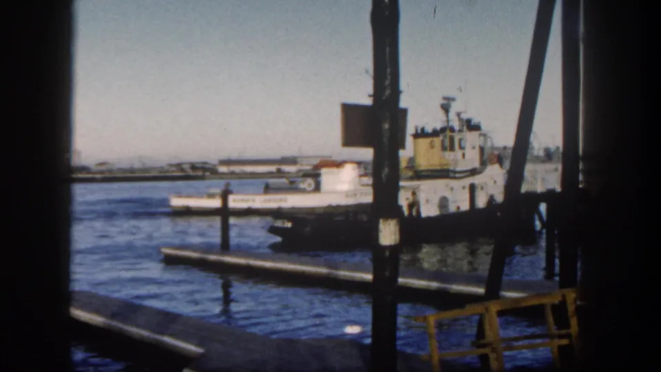 A white fishing boat remains visible near a darker vessel and dock structures, with San Diego harbor water and vertical pier posts dominating the 1962 home-movie frame.
