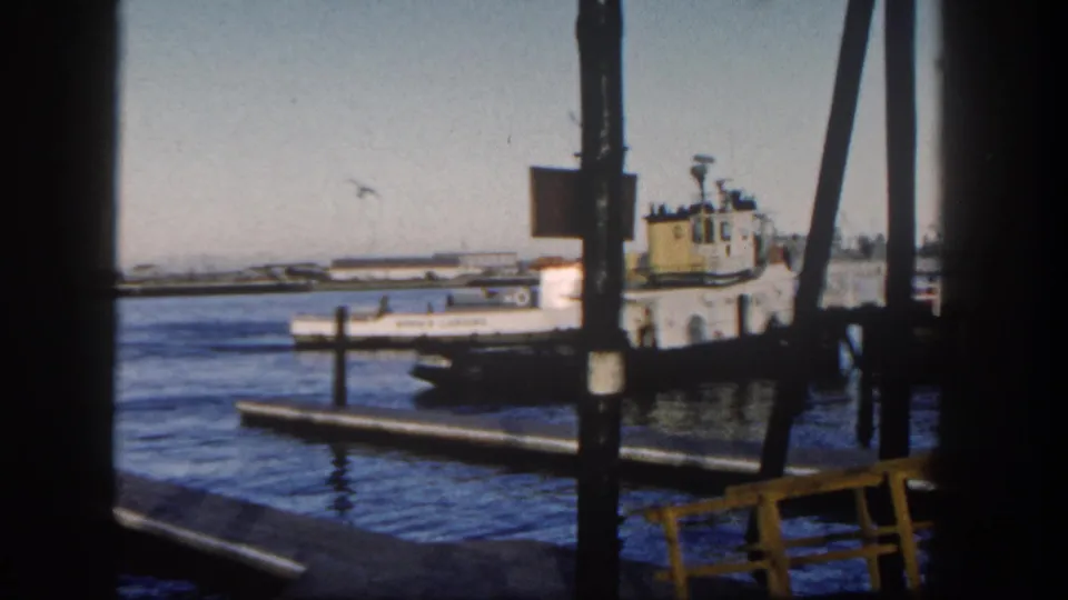 1962 San Diego Harbor view of a white and yellow tugboat near a pier, framed by dark structural beams and pilings.