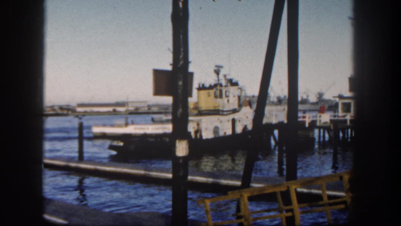 1962 San Diego Harbor tugboat at a pier, with dark beams in the foreground and industrial shoreline across the water.