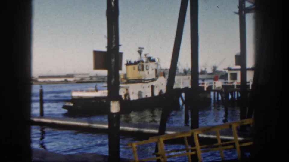 1962 San Diego Harbor scene with a tugboat, pier pilings, foreground beams, and a small dock structure to the right.