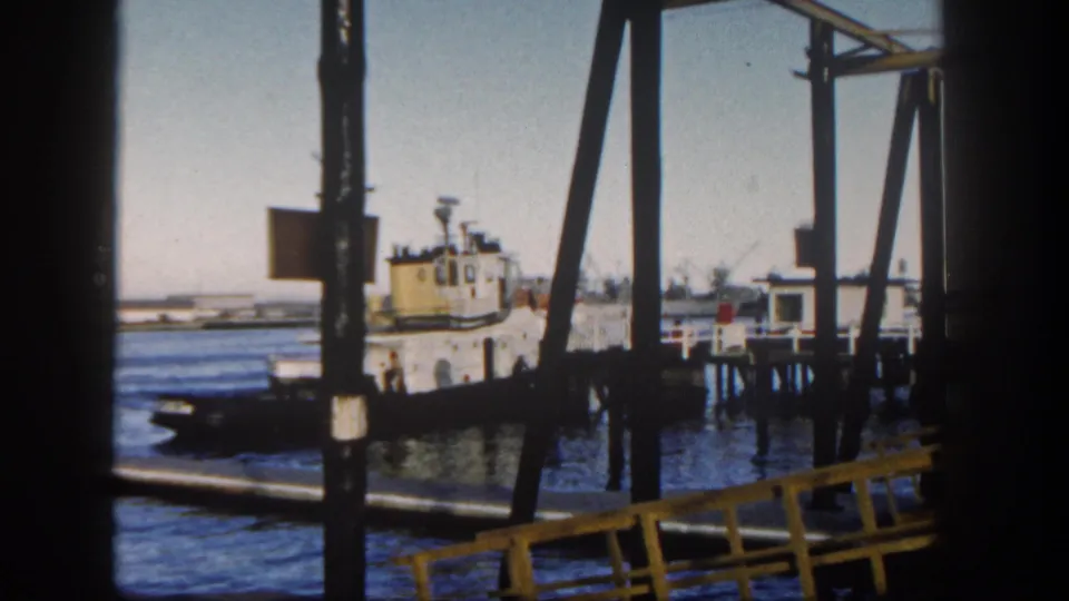 1962 San Diego Harbor tugboat framed by pier beams, with dock pilings, blue water, and a small structure at the right edge.