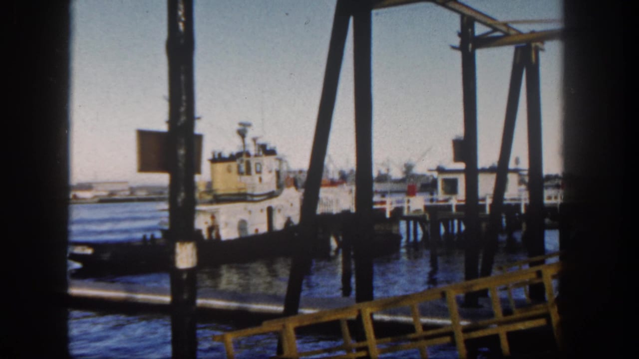 1962 San Diego harbor view framed by dark pier supports, with a small tugboat near docks and distant shoreline buildings.