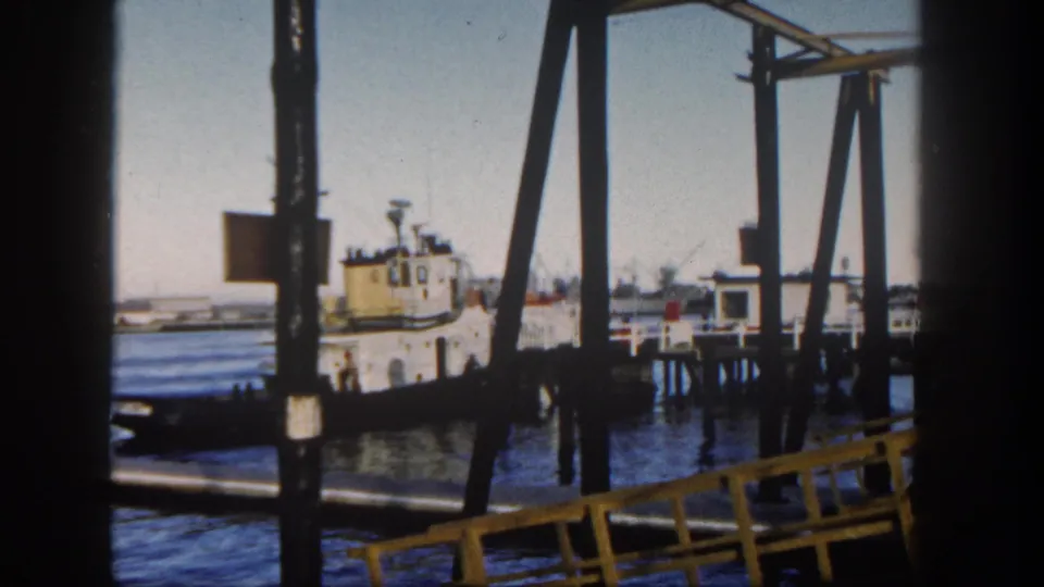 1962 San Diego harbor view framed by dark pier supports, with a small tugboat near docks and distant shoreline buildings.