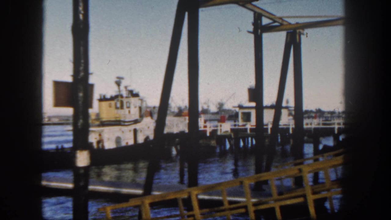 San Diego harbor scene from 1962 with dark pier beams, yellow railing, calm water, docked boats, and low buildings in the distance.