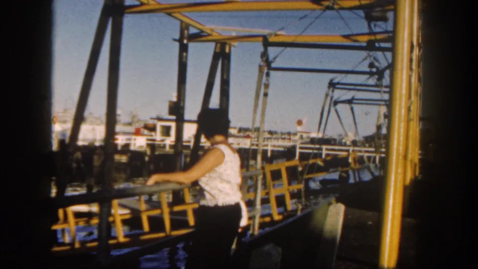 Adult woman in a white sleeveless top and dark pants stands at a yellow railing on a San Diego harbor pier in 1962.