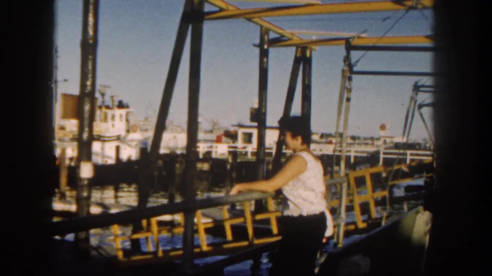 A woman stands near a yellow railing in the 1962 San Diego harbor scene, framed by dark supports, water, and distant boats.