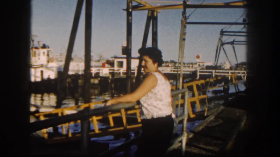 Woman on a San Diego pier in 1962 leaning on a yellow railing beside harbor water and boats.