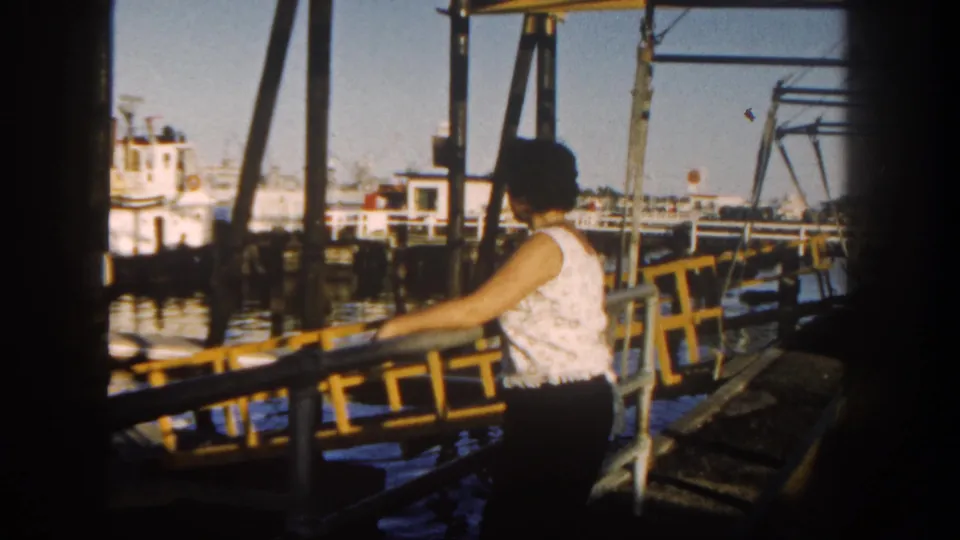 1962 San Diego harbor view with a woman at a yellow pier railing and boats in the background.