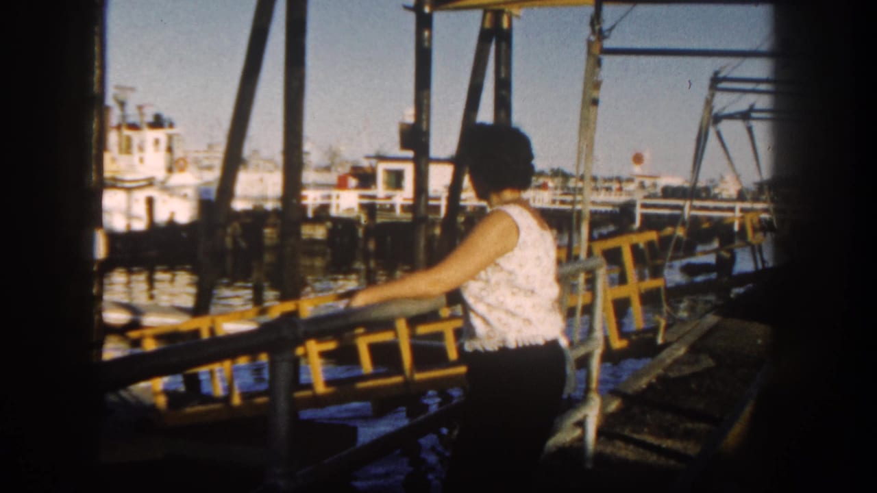 In 1962 San Diego, a woman in a white sleeveless top stands at a yellow dock railing near boats and pier supports.