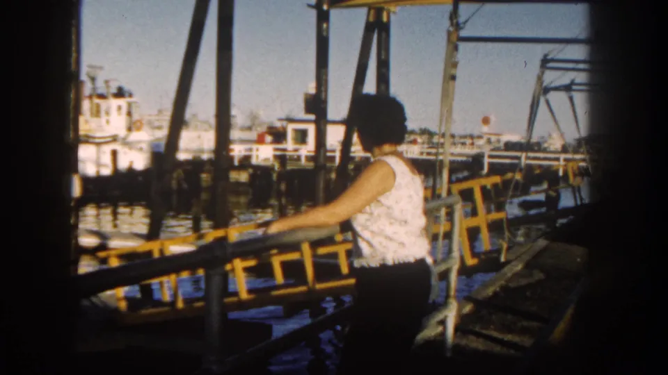 In 1962 San Diego, a woman in a white sleeveless top stands at a yellow dock railing near boats and pier supports.
