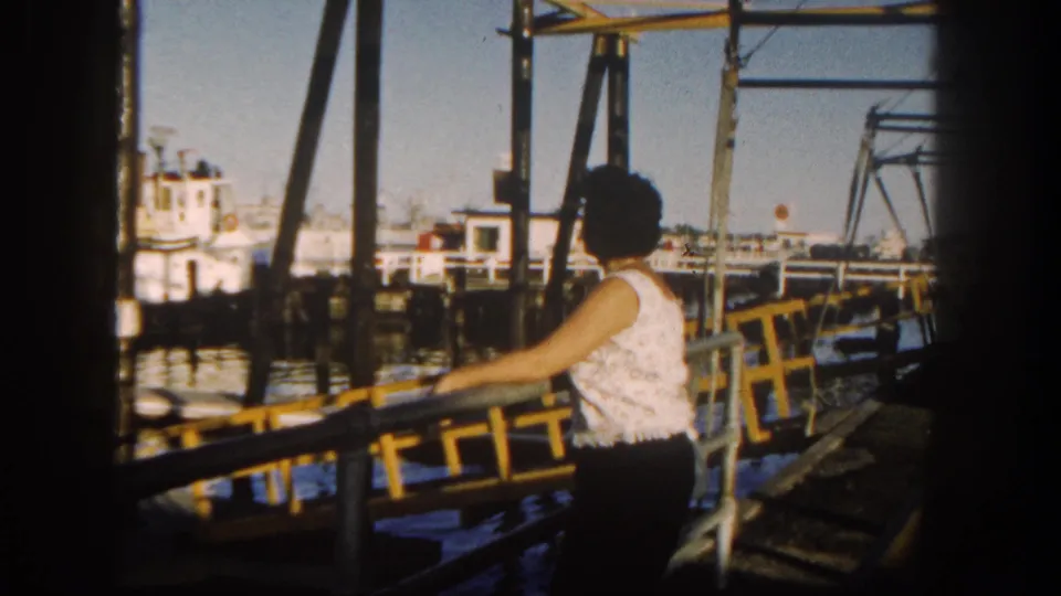 The home-movie frame shows the woman beside a yellow dock railing, with San Diego waterfront boats and pier supports behind her.