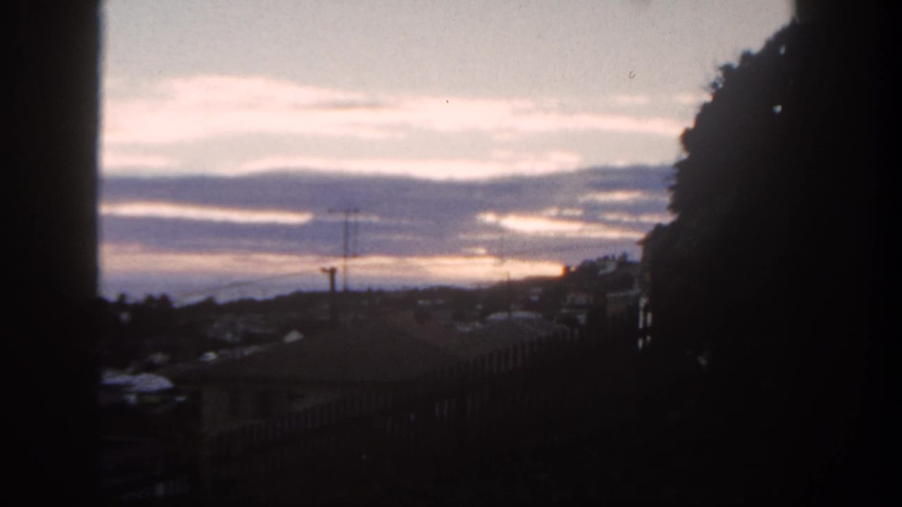 1962 San Diego suburban sunset seen through a dark window frame, with rooftops, antenna, distant hills, and clouds.