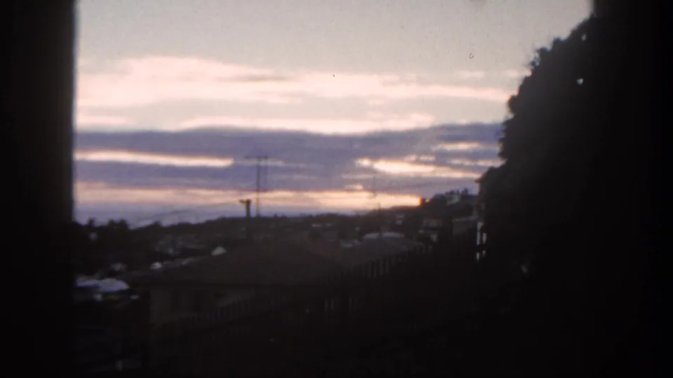 1962 San Diego suburban sunset seen through a dark window frame, with rooftops, antenna, distant hills, and clouds.