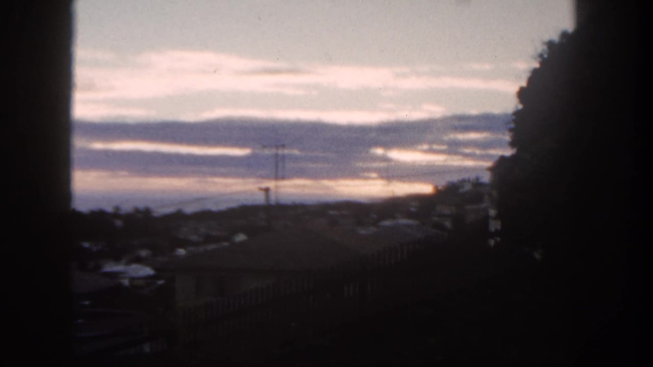 San Diego suburban hillside view at sunset in 1962, framed by a dark window edge with rooftops and an antenna visible.