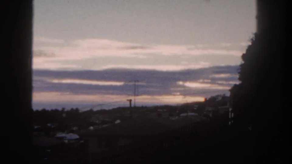 Soft 8mm view from a window over San Diego suburb rooftops and hills during a muted 1962 sunset.