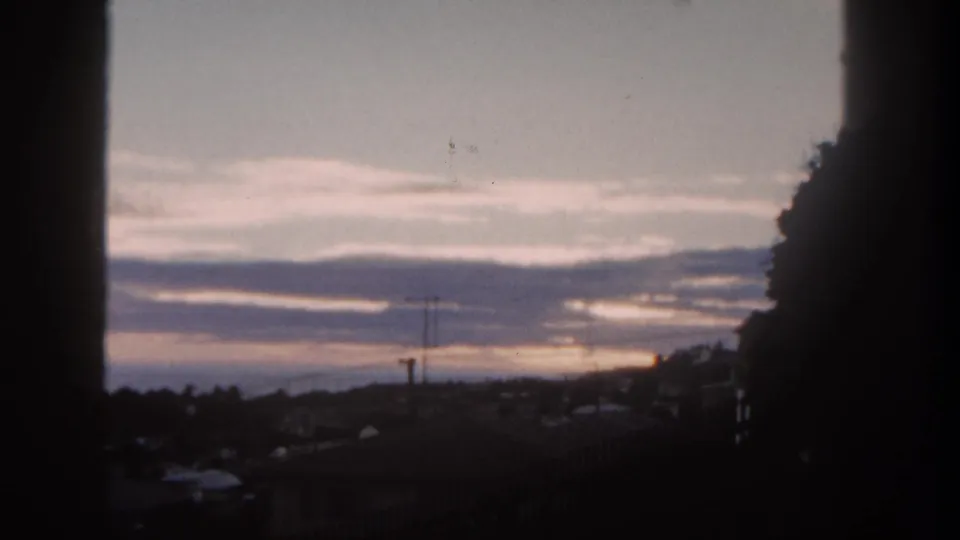 End frame of a 1962 San Diego window view, showing dark rooftops, antenna, distant hills, and purple-gray sunset clouds.