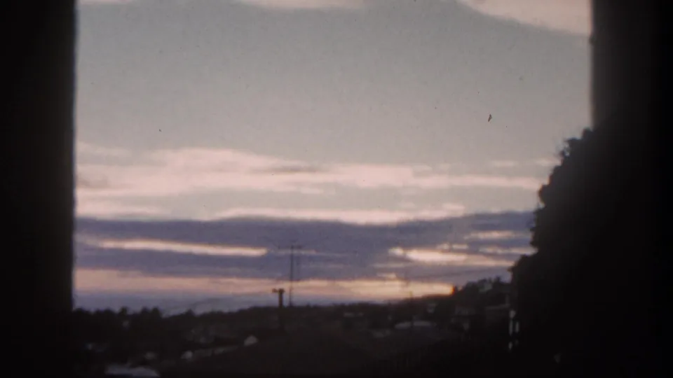 1962 San Diego home-movie frame showing a dim sunrise behind clouds, with rooftops and utility poles in silhouette.