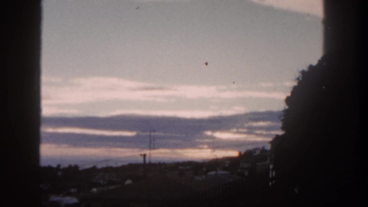 1962 San Diego frame with pale dawn light, layered clouds, utility poles, rooftops, and a dark hillside silhouette.
