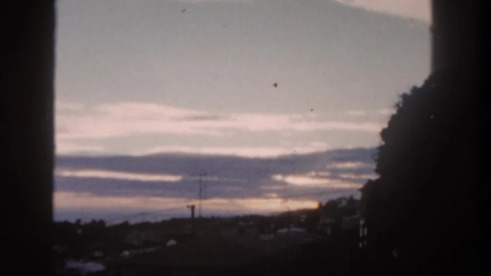 1962 San Diego frame with pale dawn light, layered clouds, utility poles, rooftops, and a dark hillside silhouette.