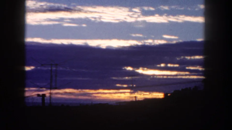 1962 San Diego 8mm frame showing orange dawn light below blue-purple clouds, with silhouetted power lines and rooftops.